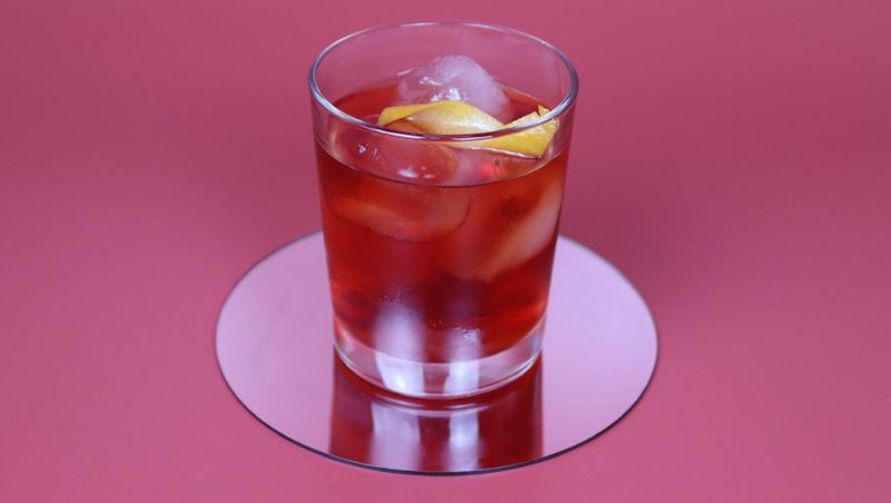 Roter Cocktail mit Eis und Orangenzeste in einem Tumbler auf einem Spiegeluntersetzer