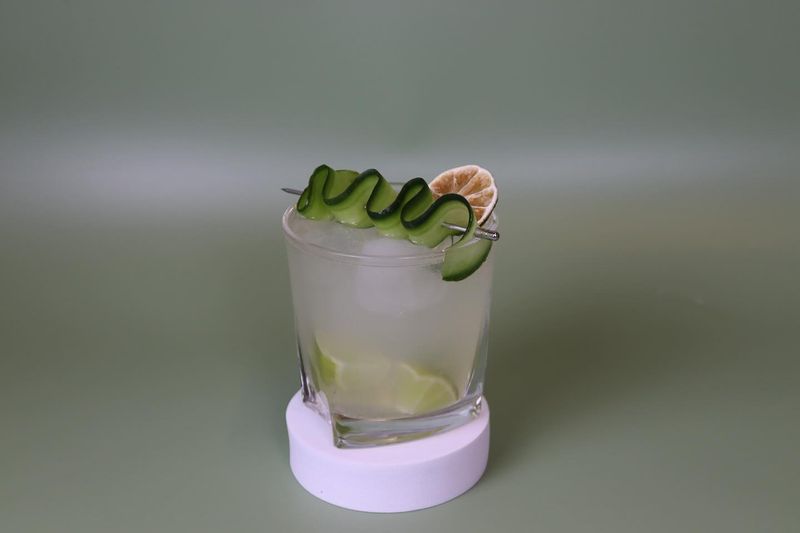 Heller Cocktail mit Gurkenspirale und Limettenscheibe auf einem fliederfarbenen Untersetzer