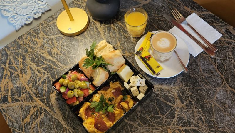 Frühstücksbento mit Gebäck, Oliven, Feta, Eierspeise und Salami, dazu ein Kaffee und Orangensaft auf einem Marmortisch