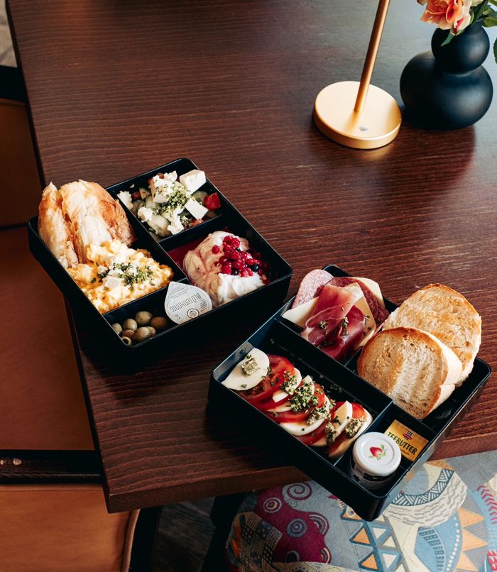 Zwei Frühstücksbentos mit Brot, Eierspeise, Oliven, Feta, Prosciutto und Tomaten-Mozzarella auf einem Holztisch