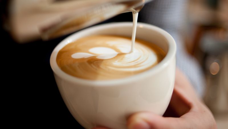 Barista gießt Milchschaum mit Latte-Art in eine weiße Kaffeetasse