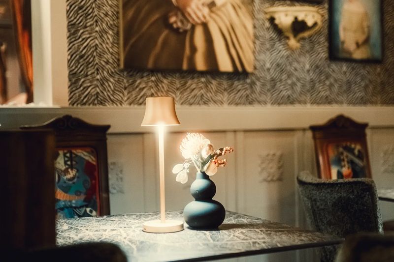 Goldene Tischlampe und schwarze Vase mit Blüte auf einem Tisch, dahinter Bilderrahmen an der Wand