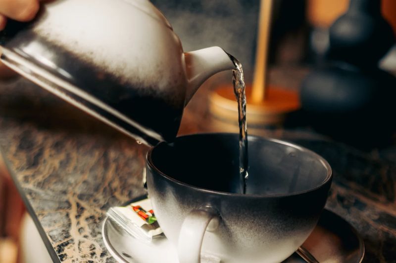 Heißes Wasser wird aus einer Kanne in eine dunkle Teeschale auf einem Marmortisch gegossen